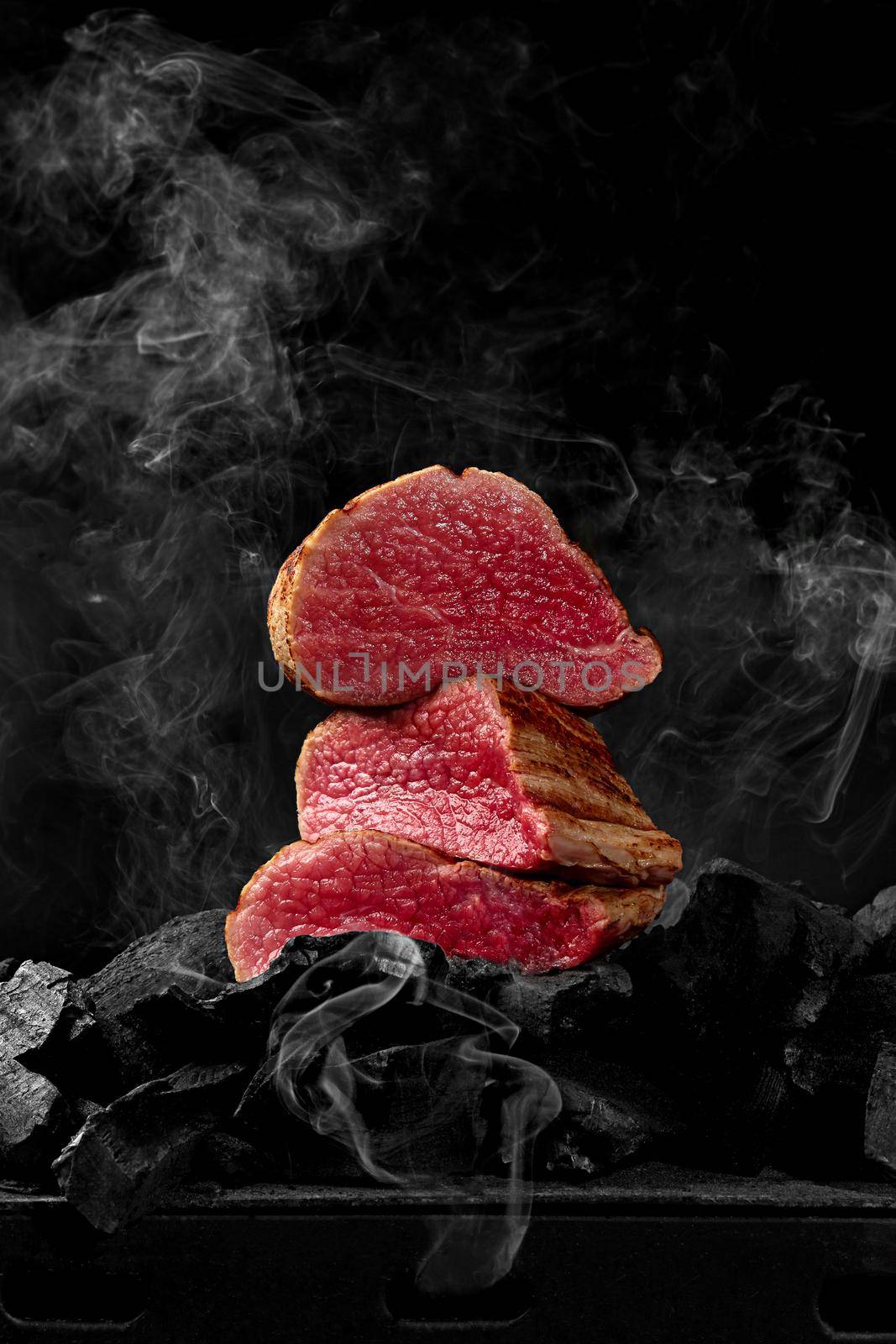 Sliced juicy rare veal tenderloin stacked on coals on bbq grill on black background in light white smoke