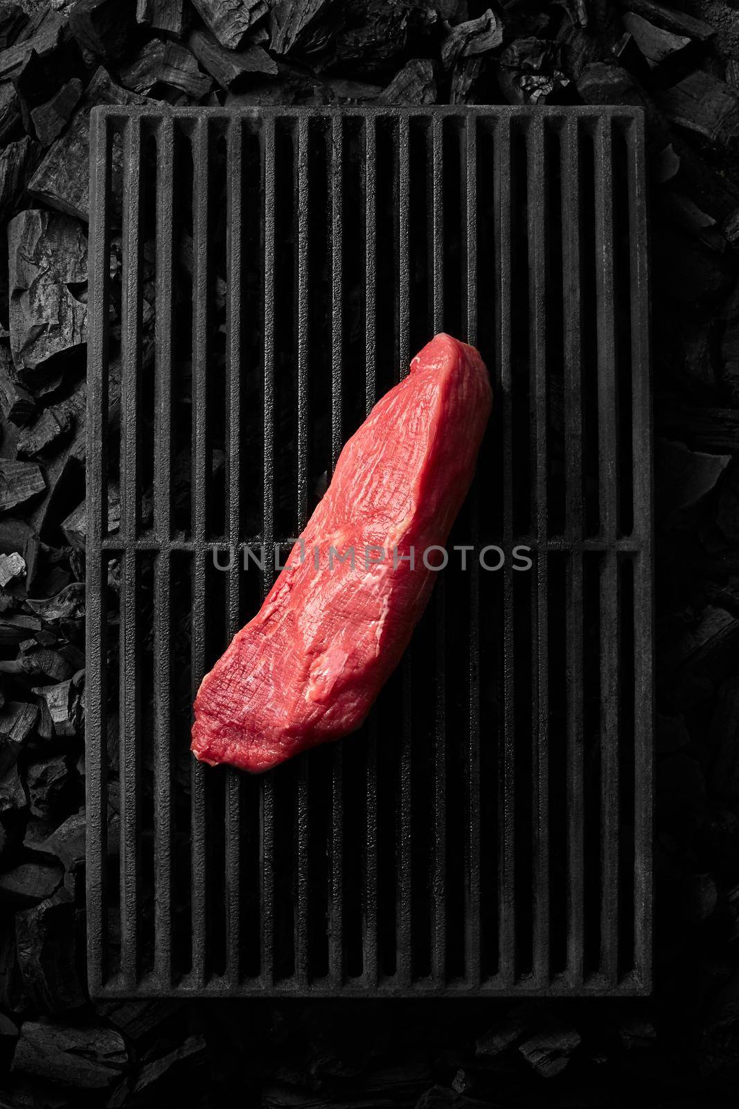 Top view of piece of lean pork tenderloin lying on black cast iron grill grate over cold coals ready for cooking