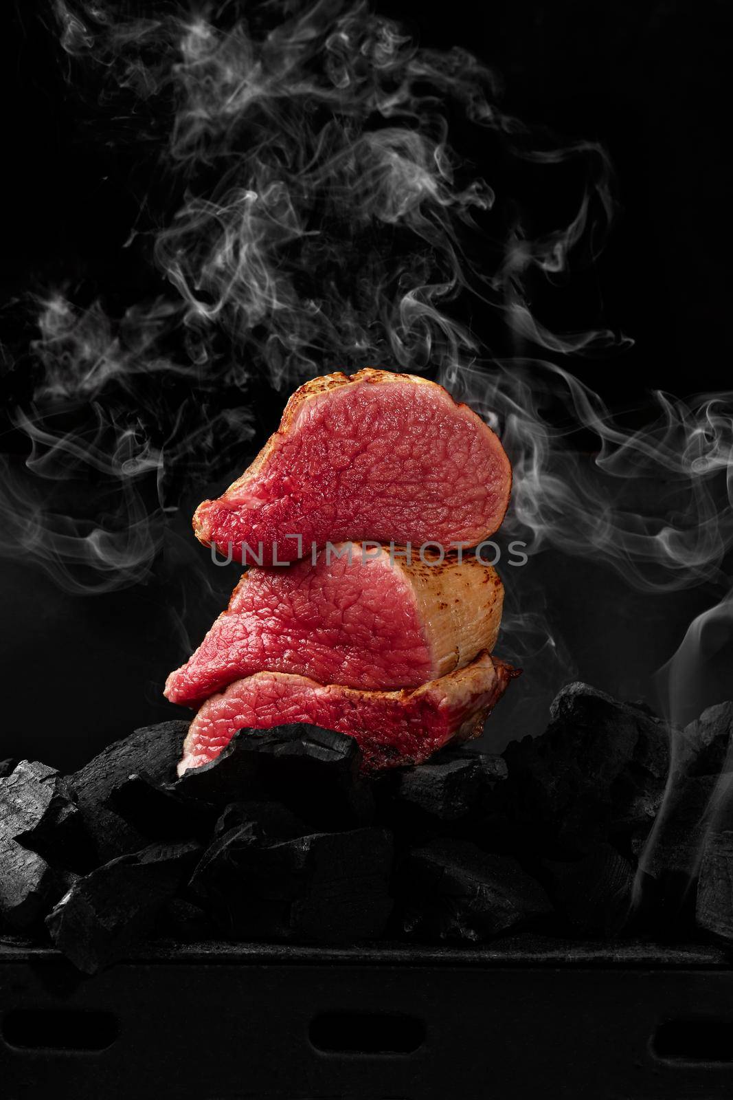 Closeup of slices of tender grilled filet mignon on charcoals on black background in light white smoke