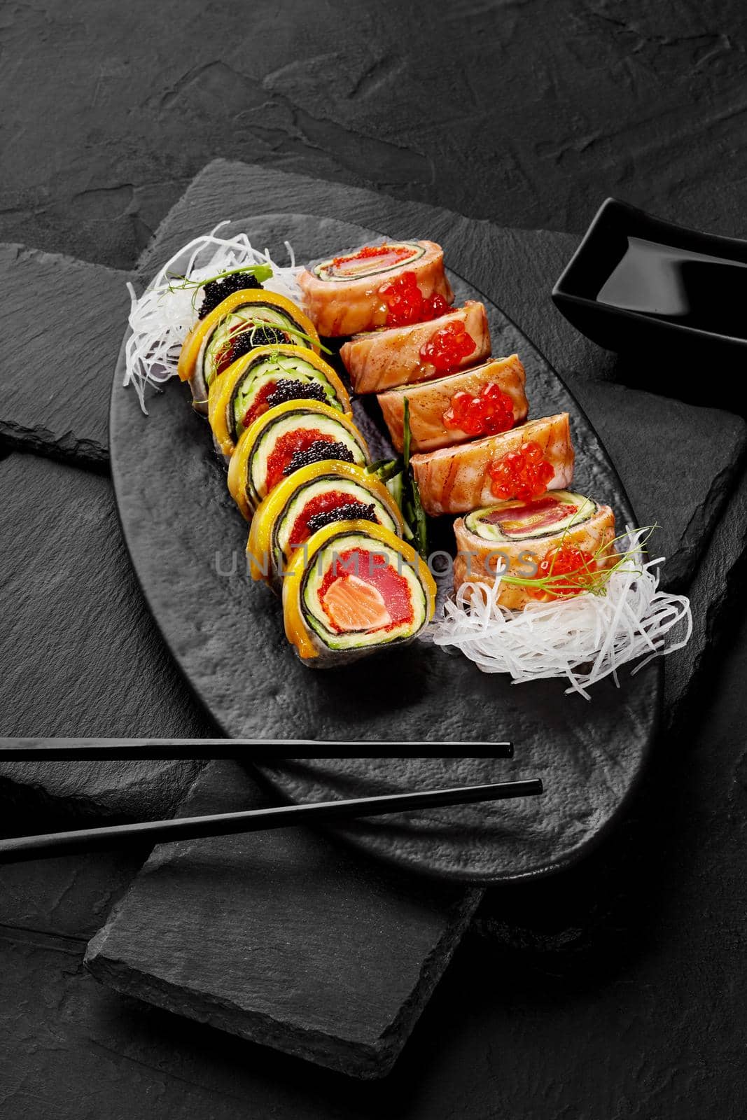 Two types of rolls with salmon, tuna, mango and avocado garnished with red caviar, tobiko and grated daikon traditionally served with soy sauce on black slate board. Japanese cuisine