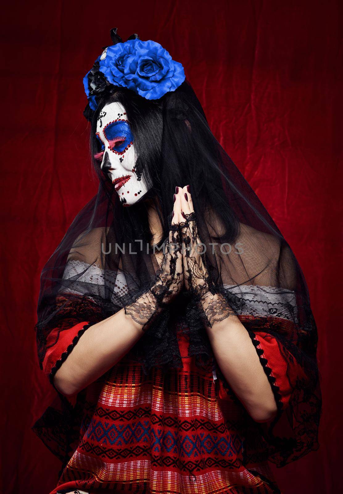 beautiful woman with a sugar skull makeup with a wreath of flowers on her head and a skull hands raised up in prayer pose in black gloves, red background by ndanko