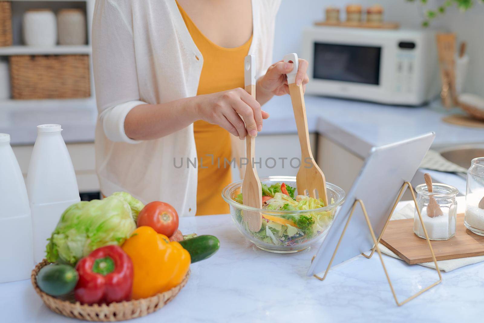 Beautiful smiling woman eating vegetables while making a salad from a recipe on the tablet computer in a kitchen by makidotvn
