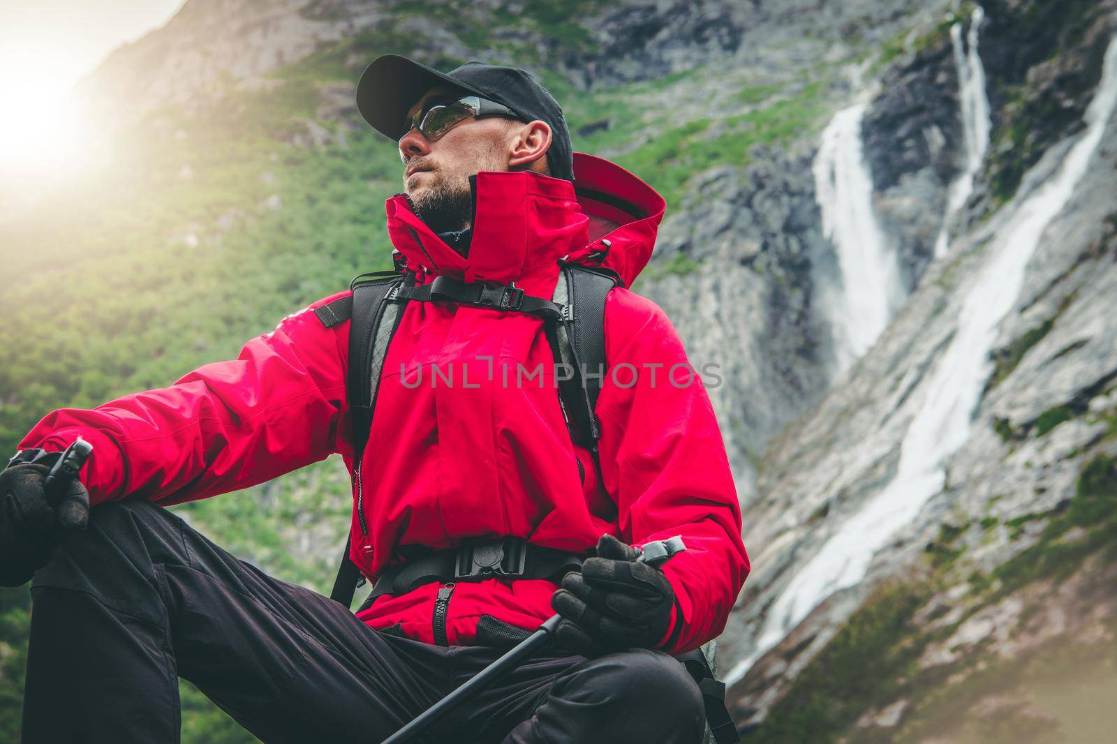 Outdoor Recreation. Active Caucasian Hiker Wearing Red Rainproof Jacket and Eyes Safety Glasses Seating Next to Scenic Alpine Waterfalls. Exploring the Trail.