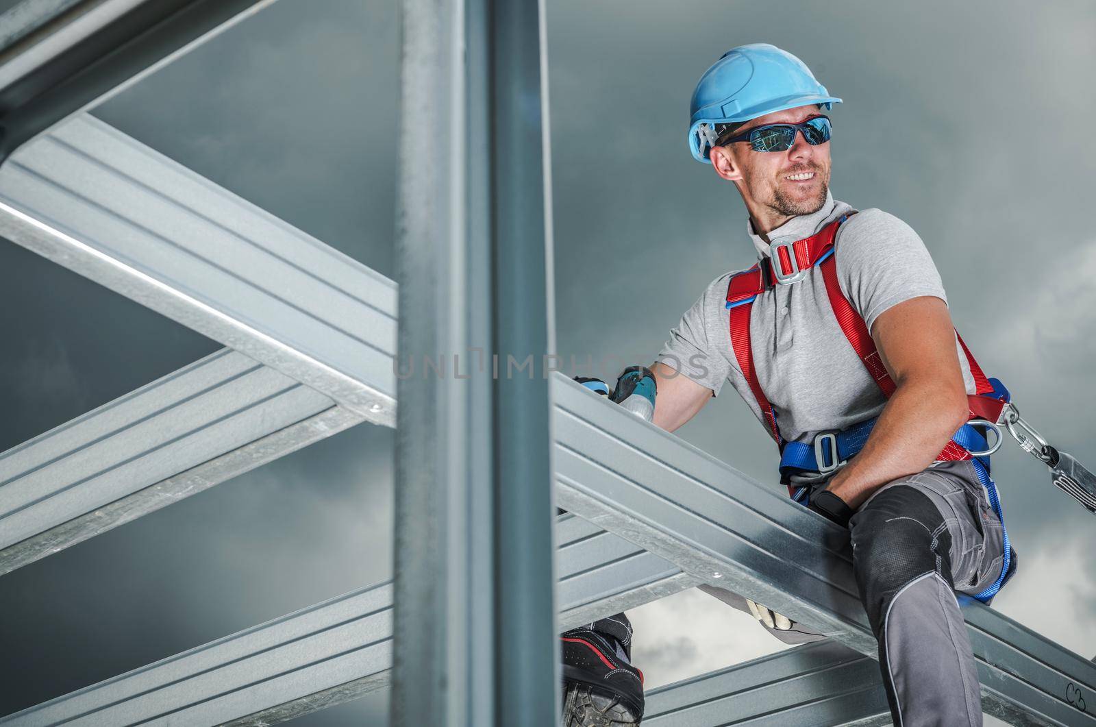 Smiling Construction Contractor Wearing Safety Harness. Caucasian Worker in His 30s. Skeleton Steel Building Frame. by welcomia