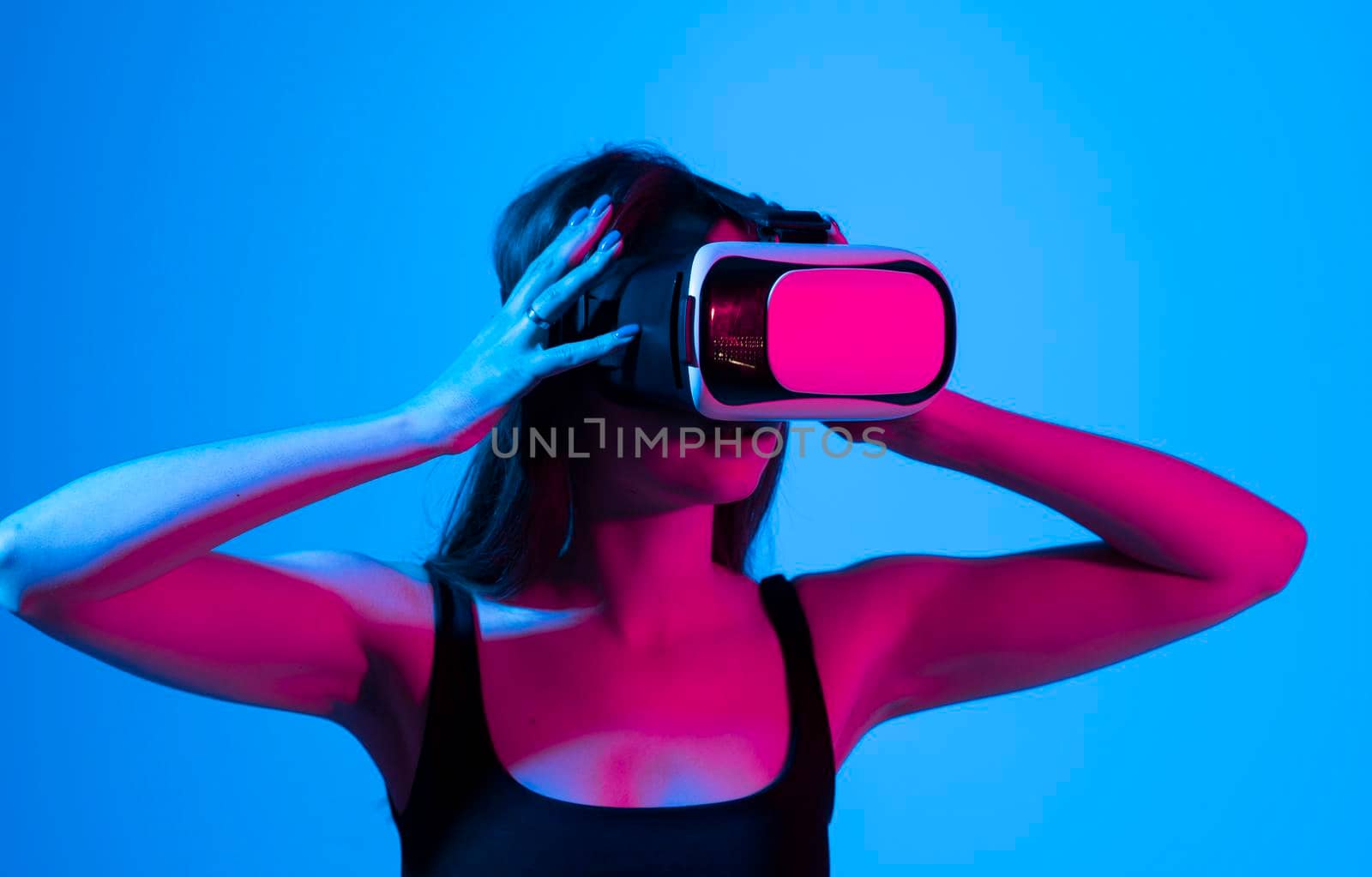 Cheerful girl with hands up wearing the virtual reality goggles and playing a games in neon light. Future technology concept. Metaverse. by vovsht