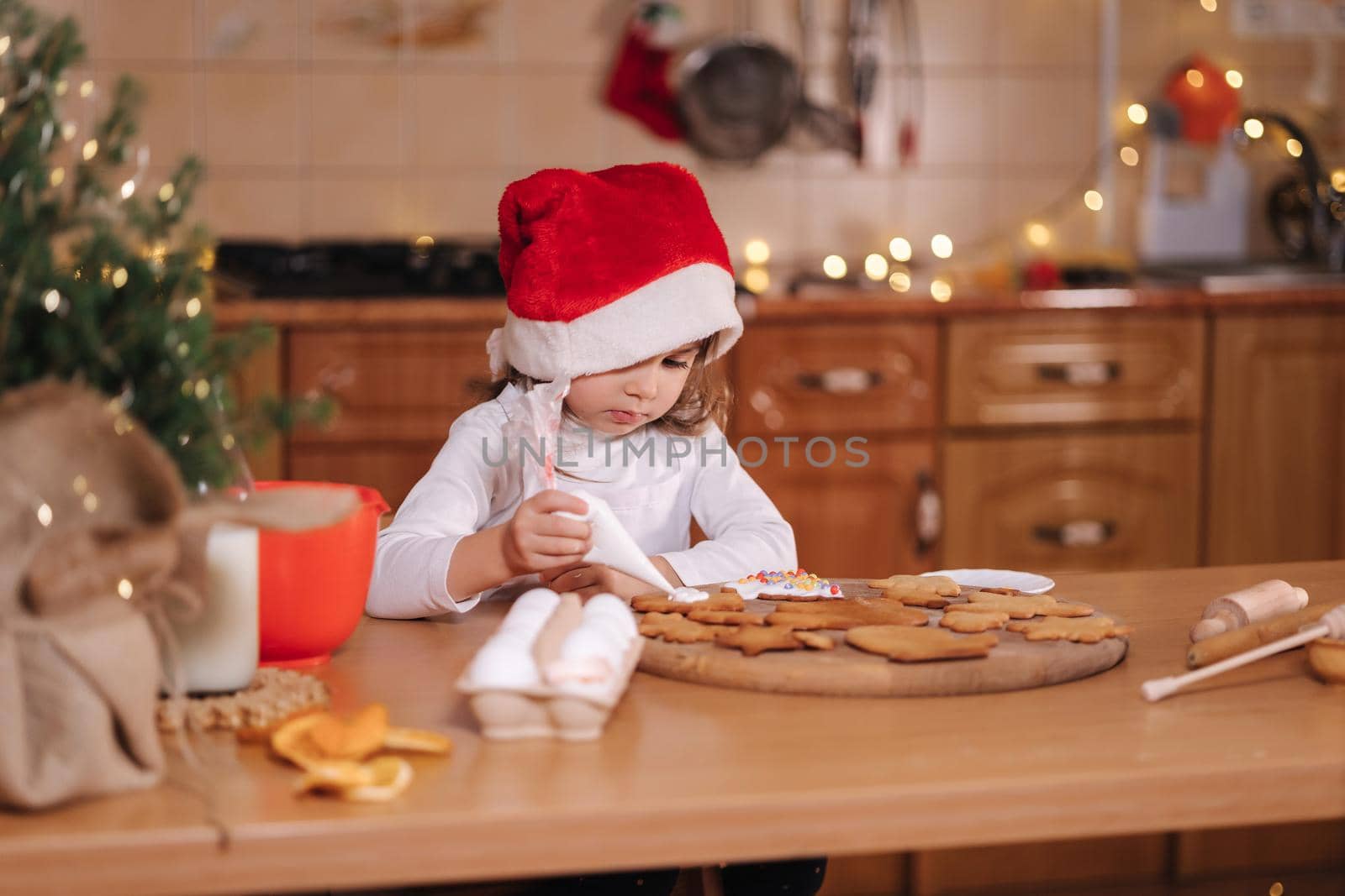 Little girl in santa hat decorates gingerbread using multicolored beads. Christmas and New Year traditions concept. Christmas bakery. Happy hollidays.