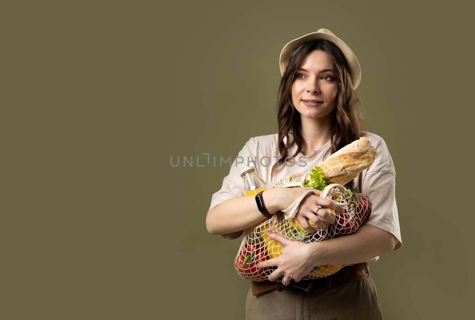 Smiling young woman in light summer clothes with a mesh eco bag full of vegetables, greens watching in a camera on a green studio background. Sustainable lifestyle. Eco friendly concept. Zero waste. by vovsht