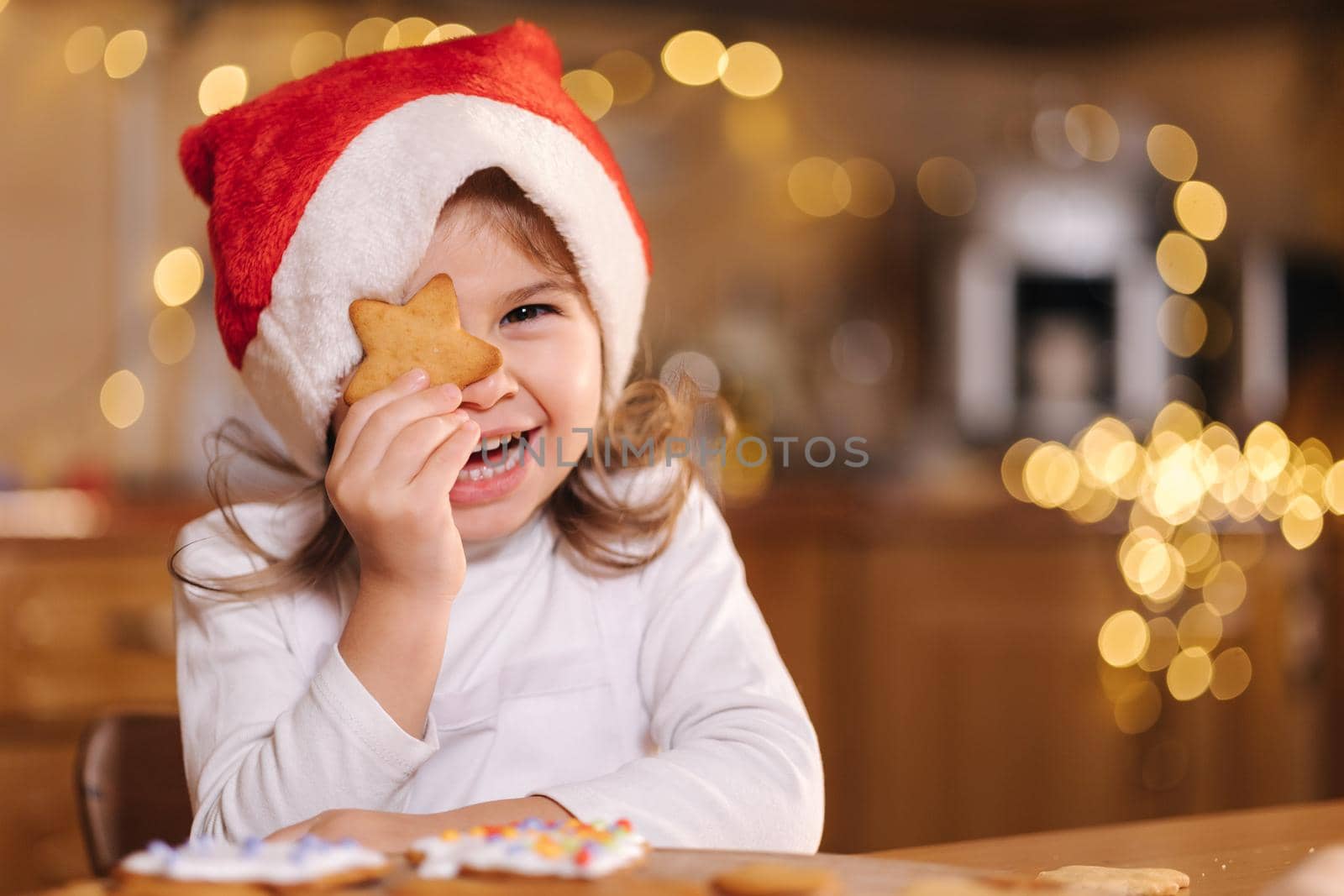 Adorable little girl in santa hat close one eye by gingerbread and laughing. Happy little kid preparing for Christmas. Background of fairy lights bokeh.