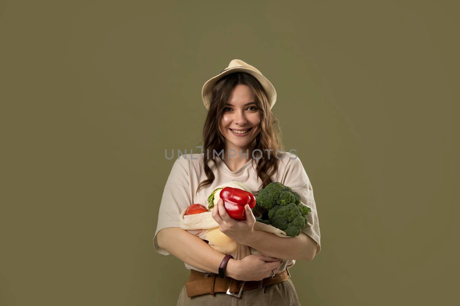 Portrait of happy smiling young woman in beige oversize t-shirt holding reusable string bag with groceries over green background. Sustainability, eco living and people concept. by vovsht