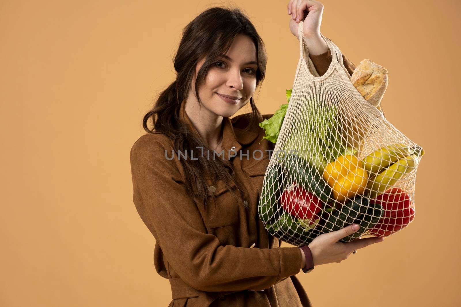 Portrait of happy smiling young woman in brown dress holding reusable string bag with groceries over orange background. Sustainability, eco living and people concept