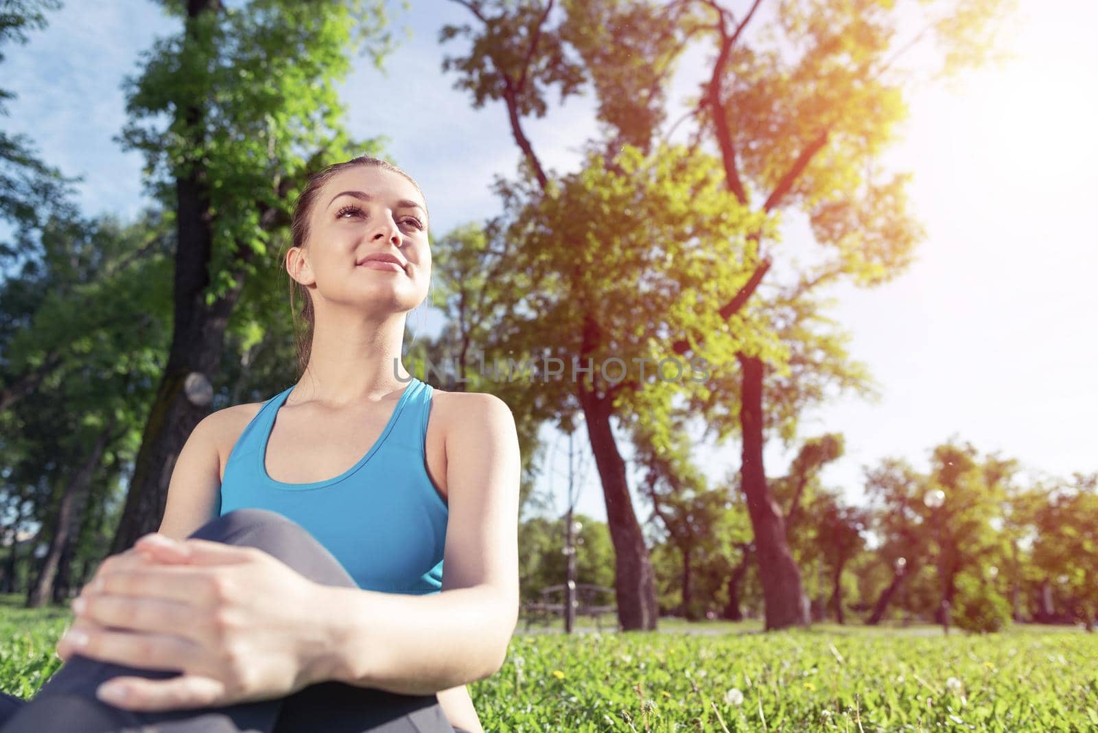 Beautiful smiling girl in sportswear relax in park. Portrait of young charming woman sitting on green grass. Training and meditation outdoor at sunny day. Morning exercises and healthy lifestyle.