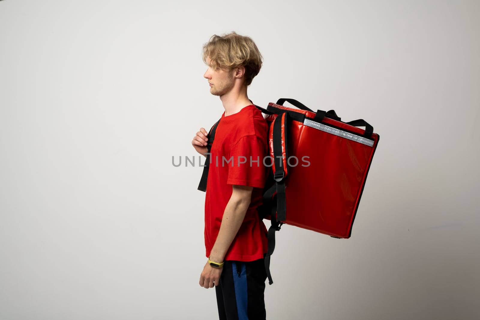 Young delivery employee man in red tshirt uniform with a thermal food bag backpack works as a courier in a food delivery service on a white background. by vovsht