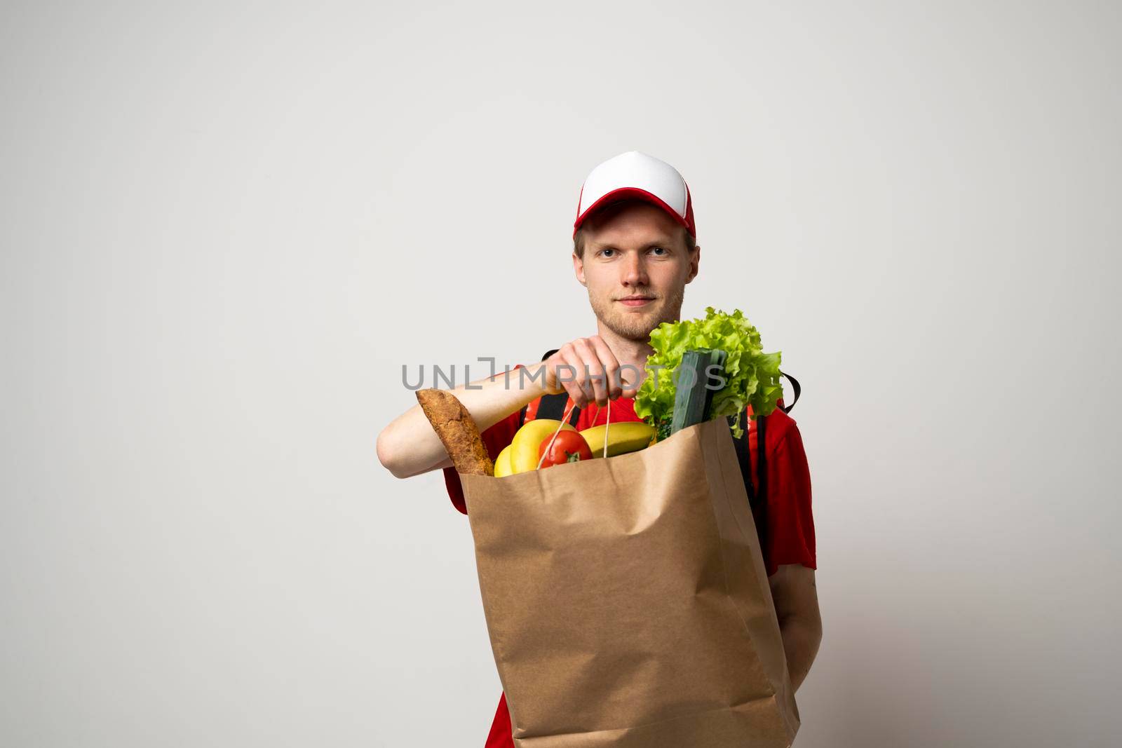 Delivery man employee in red cap and t-shirt uniform hold craft paper packet with food isolated on white background studio. by vovsht