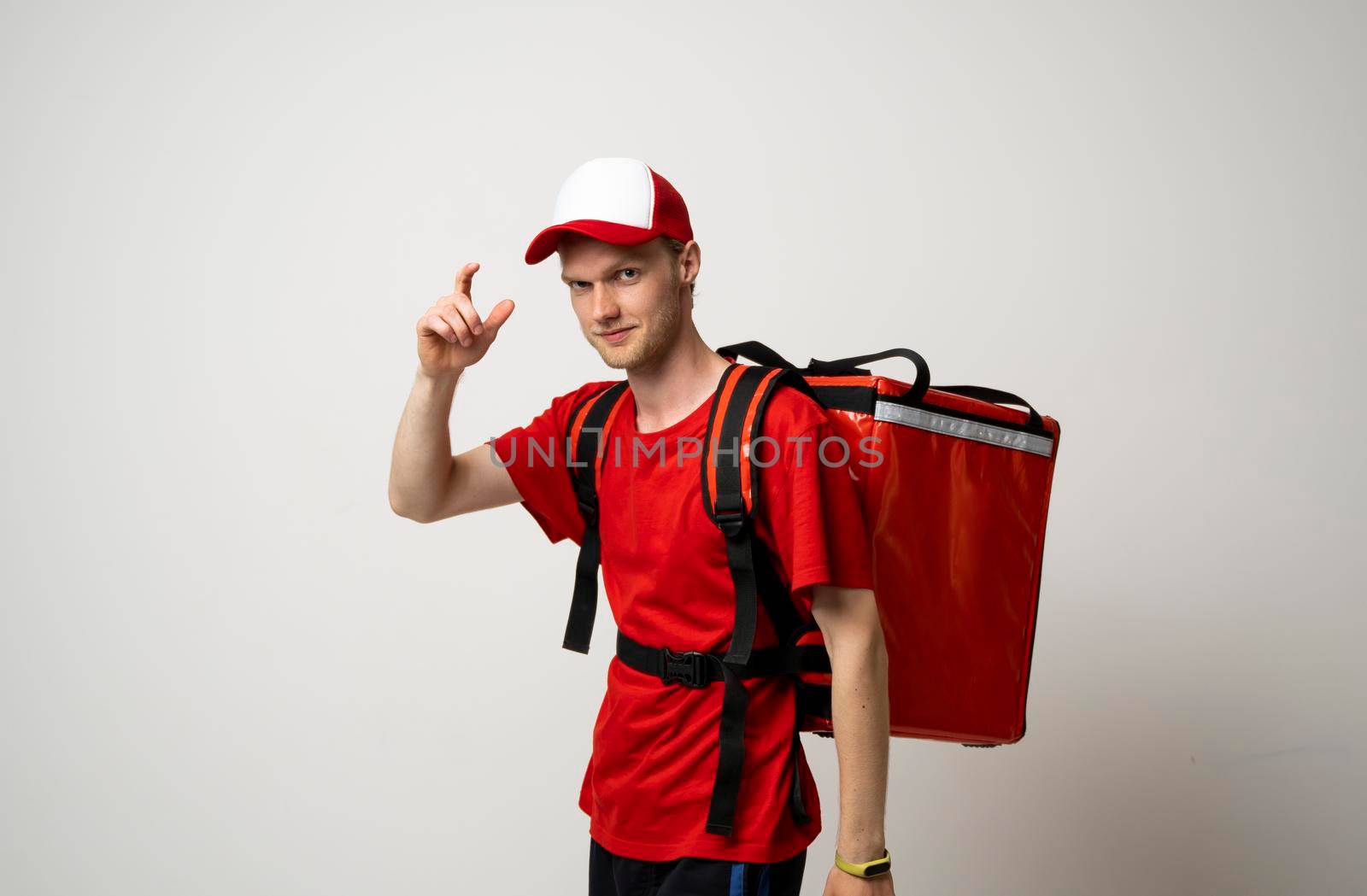Young courier with thermal bag on color background, space for text. Food delivery service. Delivery guy in a red t-shirt uniform work as courier and holds red thermal food backpack. Service concept. by vovsht