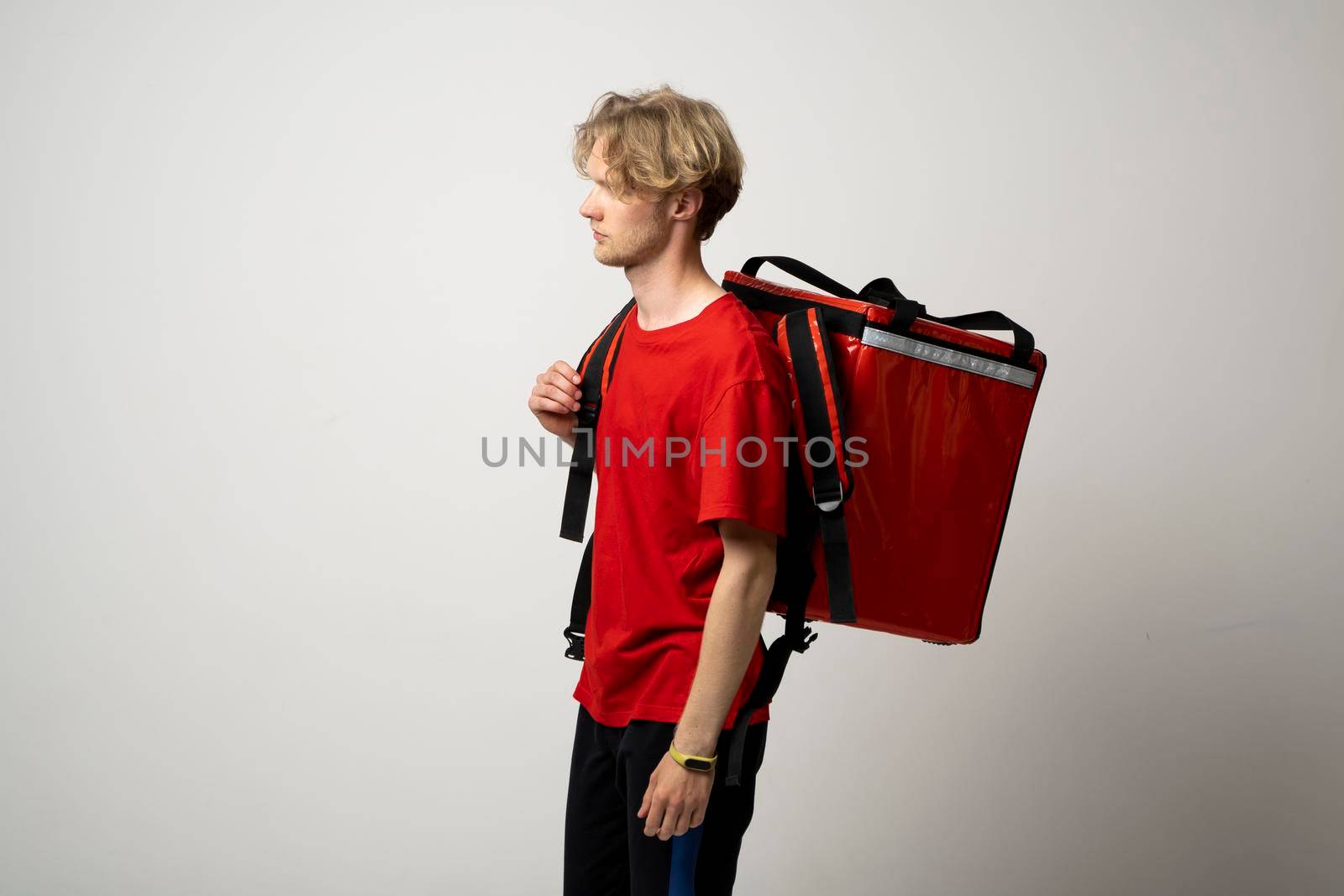 Portrait of a young courier, delivery man in red uniform with a thermal backpack isolated on a white background. Fast home delivery. Online order. Courier delivers groceries home