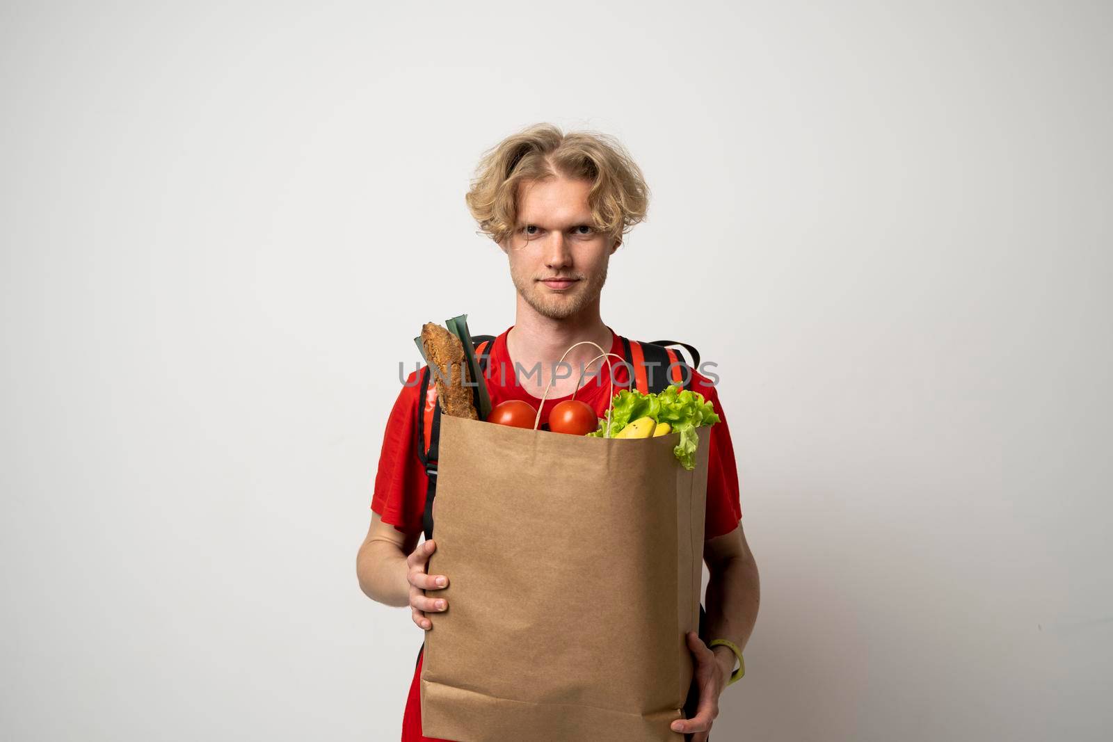 Delivery man employee in red t-shirt uniform hold craft paper packet with food isolated on white background studio. by vovsht