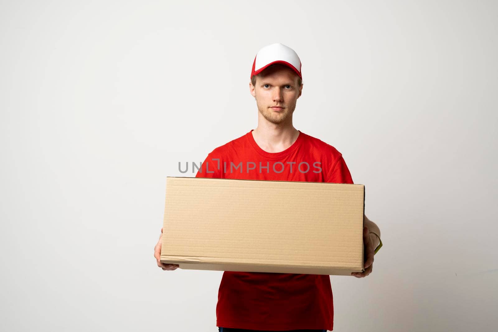Delivery service. Young smiling courier holding cardboard box. Happy young delivery man in cap and red t-shirt standing with parcel isolated on white background