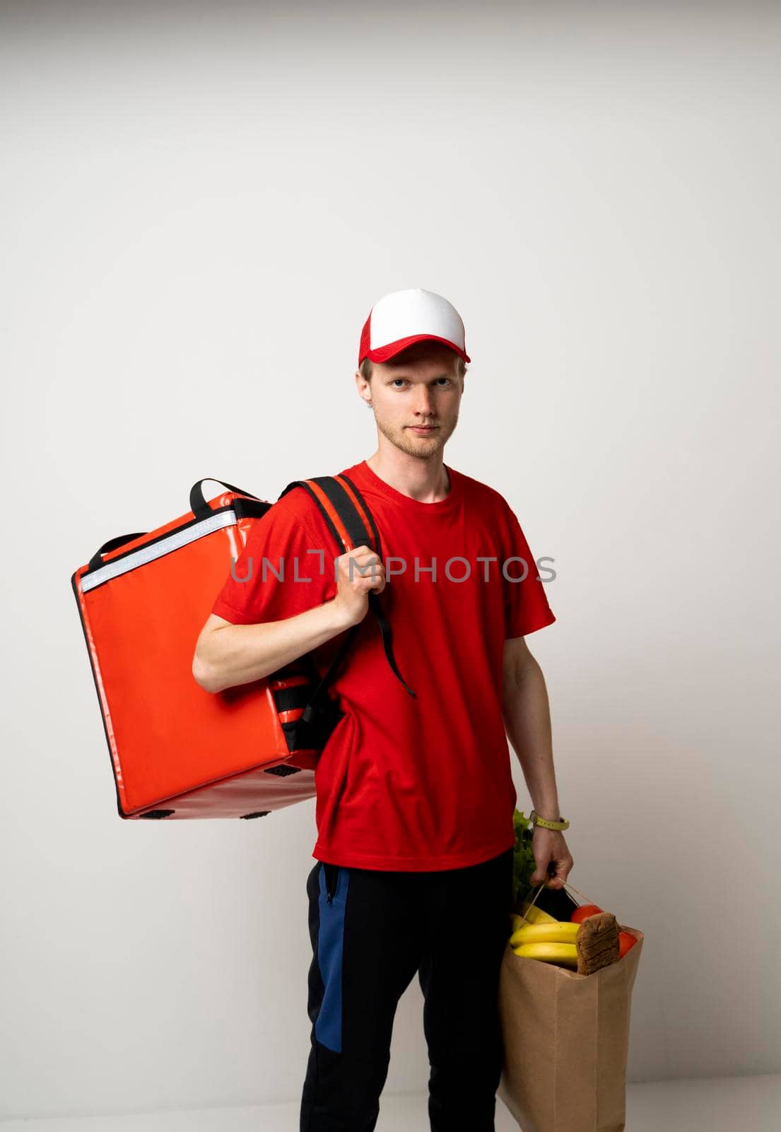 Delivery man in red uniform with a thermal bag on a shoulder holding paper bag with groceries. Food delivery service. by vovsht