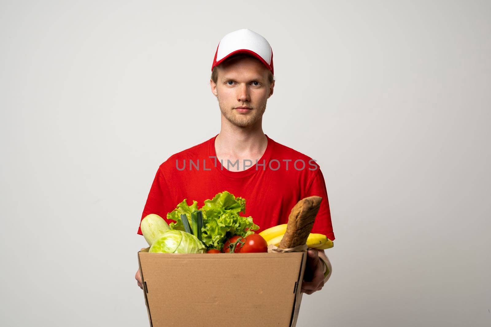 Delivery man in red uniform holds craft paper packet with food isolated on white background, studio portrait. Service concept. by vovsht