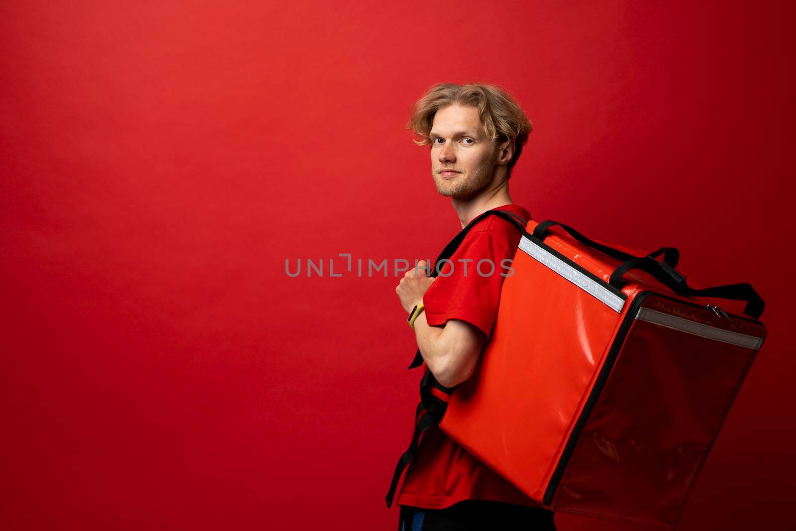 Young courier with thermal bag on red background, space for text. Food delivery service. Delivery guy in a red t-shirt uniform work as courier and holds red thermal food backpack. Service concept