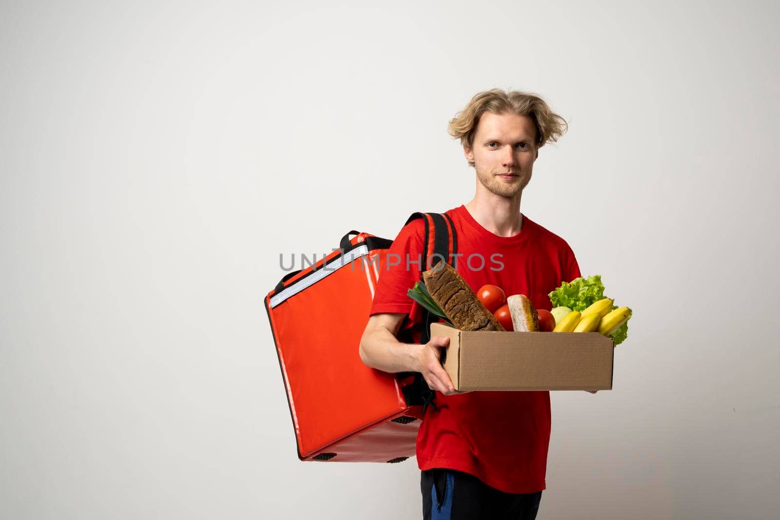Food delivery service. Portrait of pleased delivery man in red uniform smiling while carrying paper box with food products isolated over white background
