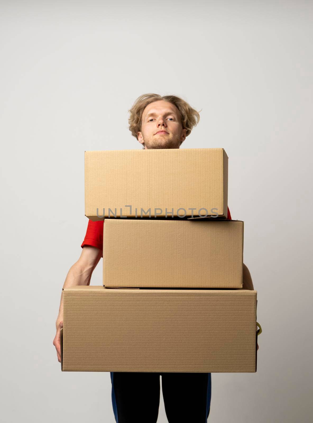 Delivery man in a red uniform holding a stack of cardboard boxes. Courier delivering postal packages over white studio background