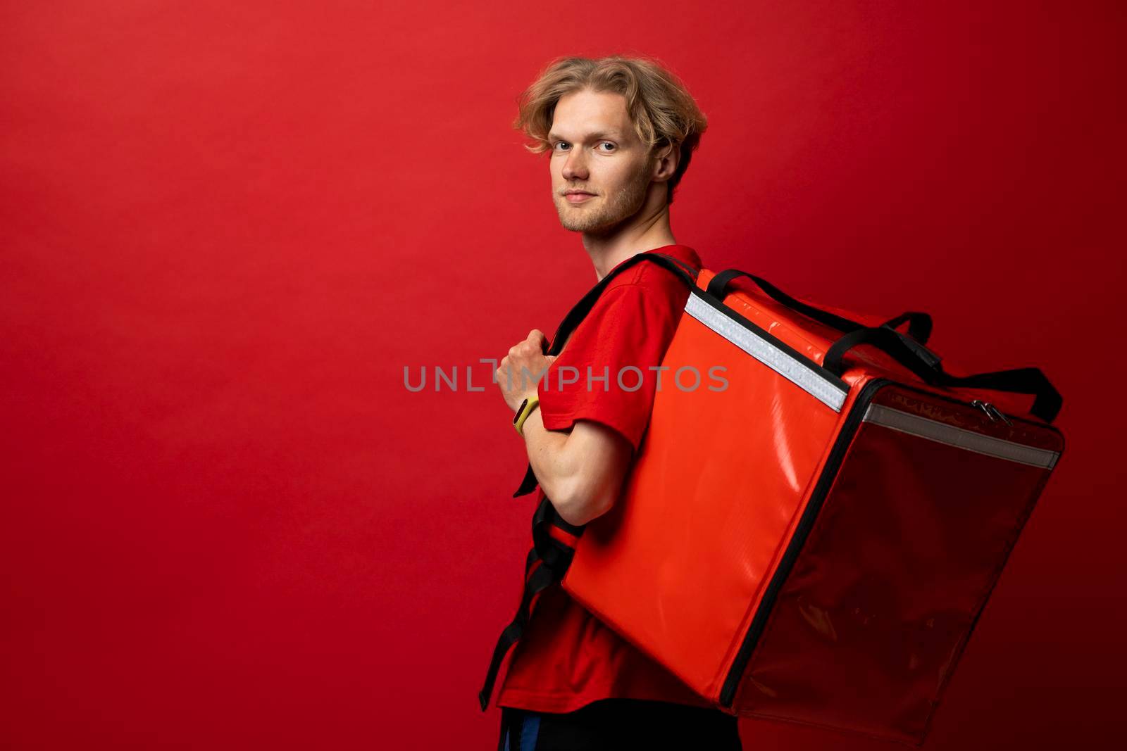 Delivery guy employee man in red cap and t-shirt uniform workwear work as dealer courier hold red thermal food bag backpack isolated on red color background studio. Service concept