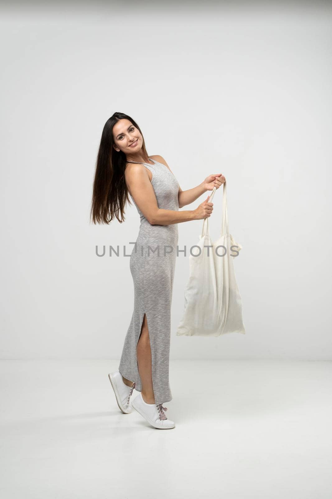 Brunette woman in a grey dress holding cotton shopper bag with groceries over white background. Rejection of plastic. Zero waste concept