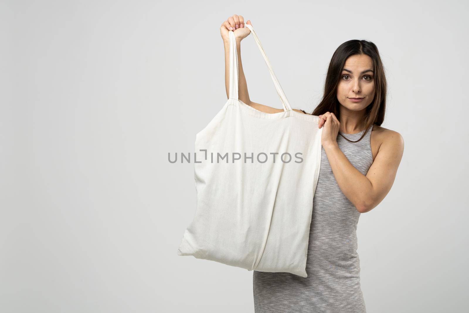 Cheerful millennial woman showing white eco bag to camera standing over white studio background. Lady holding cotton shopper handbag. fashion and ecology concept