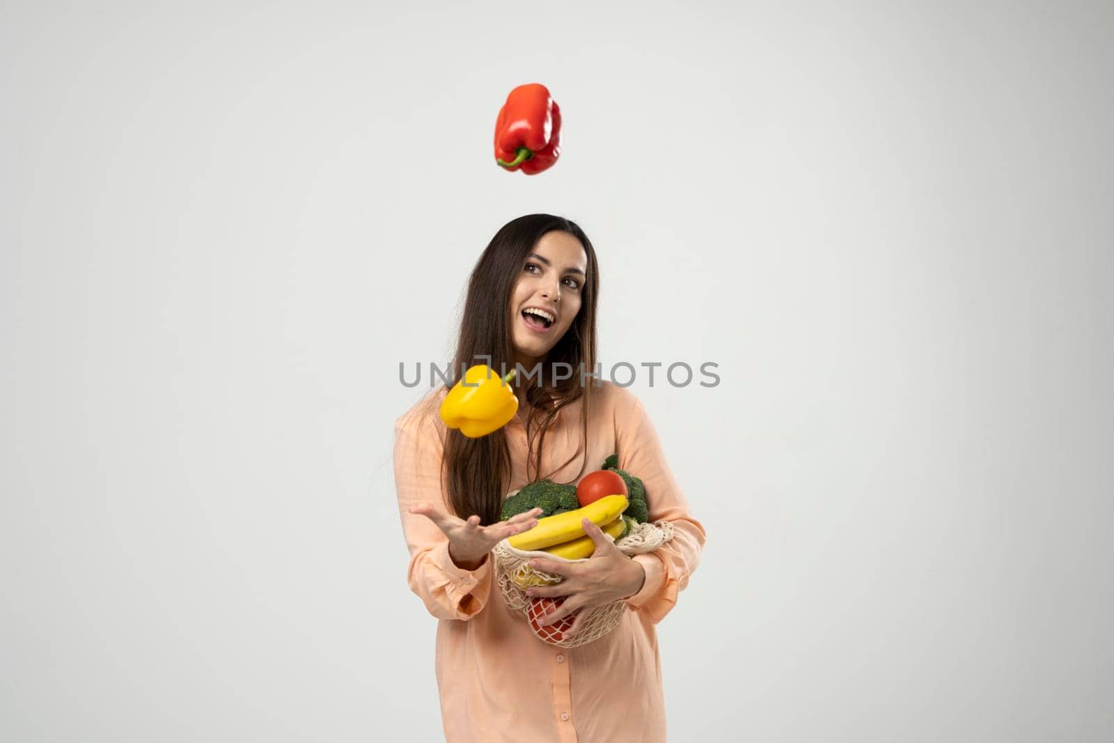 Smiling woman in dress throw up a red and yellow pepper in a air by one hand and holding reusable cotton eco bags in other