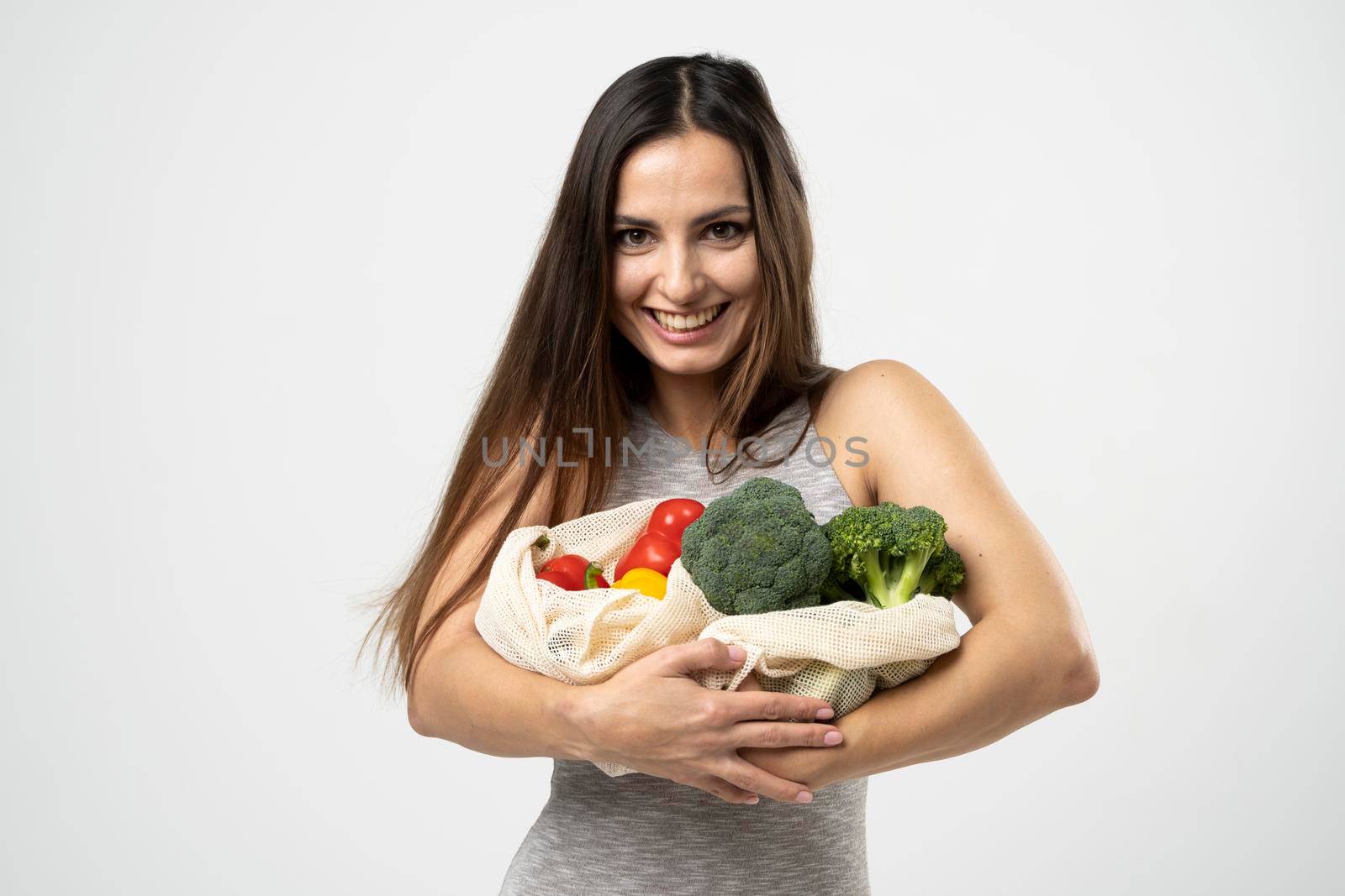 A white mesh mesh bag with groceries in a hands of a girl in grey dress against the white background of a studio. by vovsht