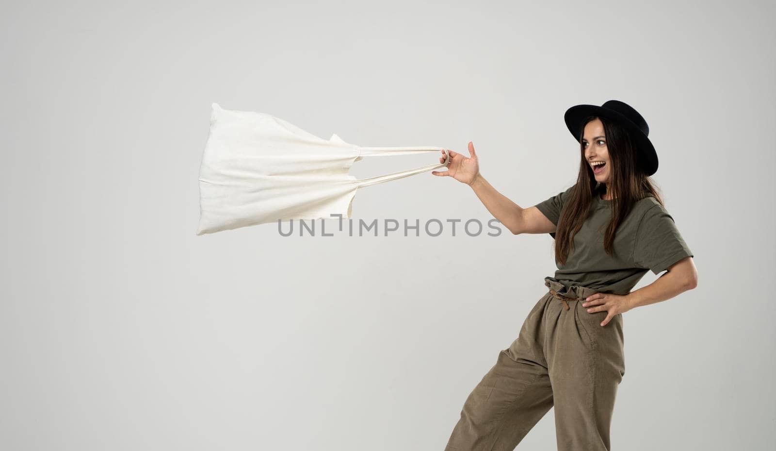 Happy young brunette woman in a green t-shirt and black hat dancing with a cotton bag with groceries. Zero waste concept. Eco friendly lifestyle. Mockup