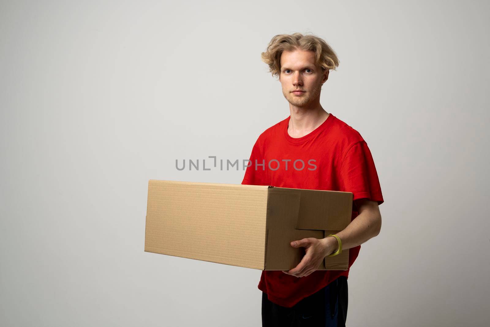 Happy young delivery man in red uniform carry cardboard box in hands on white background. Delivery guy give parcel shipment