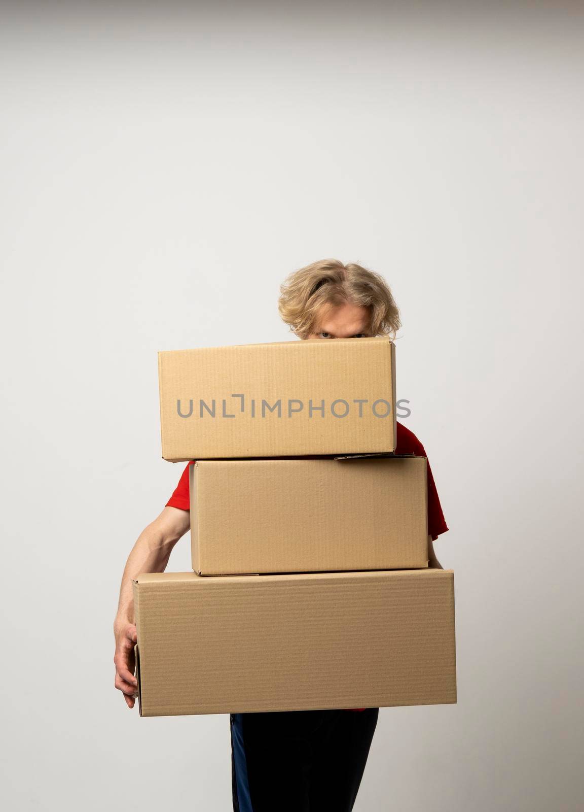 Delivery man in a red uniform holding a stack of cardboard boxes. Courier delivering postal packages over white studio background