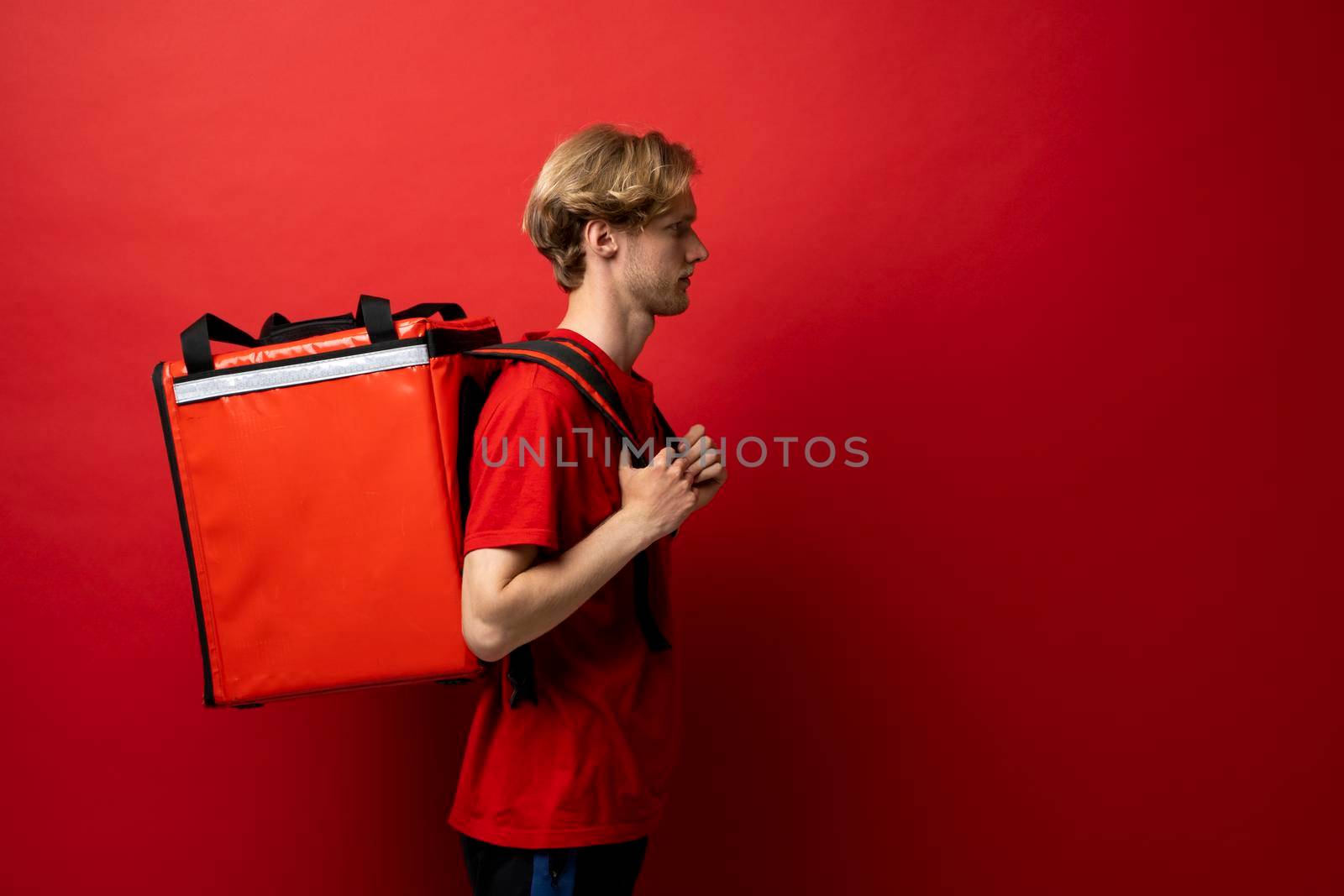 Side view of young courier in a red t-shirt and with a thermo bag on color red background, space for text. Food delivery service. by vovsht