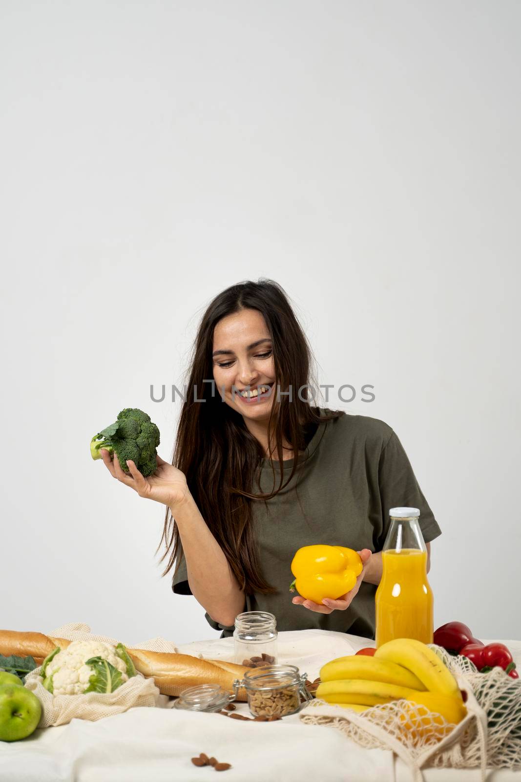 Happy woman in green t-shirt unpacking shopping mesh eco bag with healthy vegan vegetables, fruits, bread, snacks and holding broccoli and yellow pepper in a hands. Healthy eating vegetarian concept. by vovsht