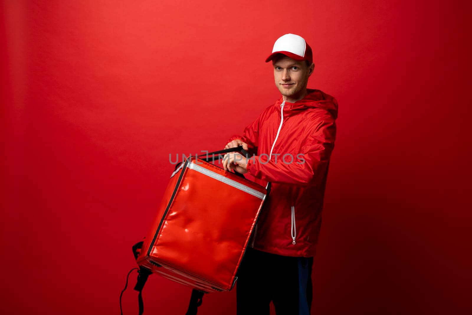 Delivery employee man 20s in red uniform with a thermal food bag backpack works in a courier service isolated on red background studio