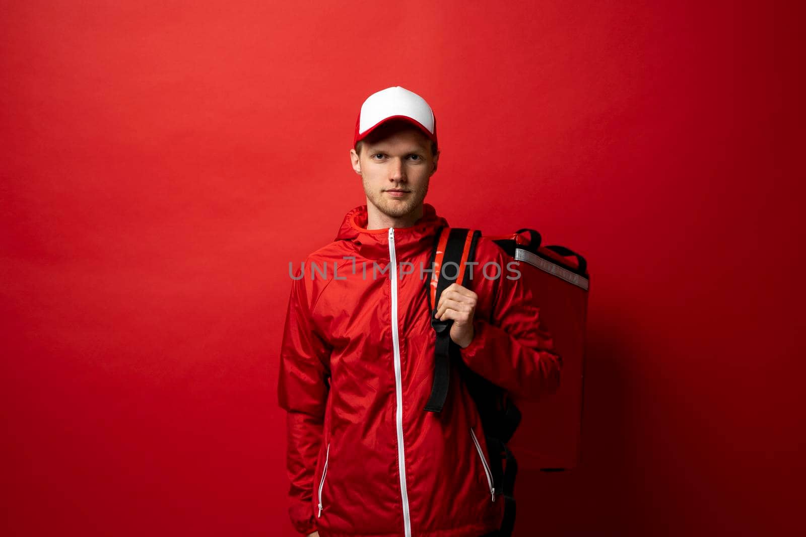 Young courier in a red uniform t-shirt and with red food thermo bag on a shoulder standing isolated on red background studio. Food delivery service