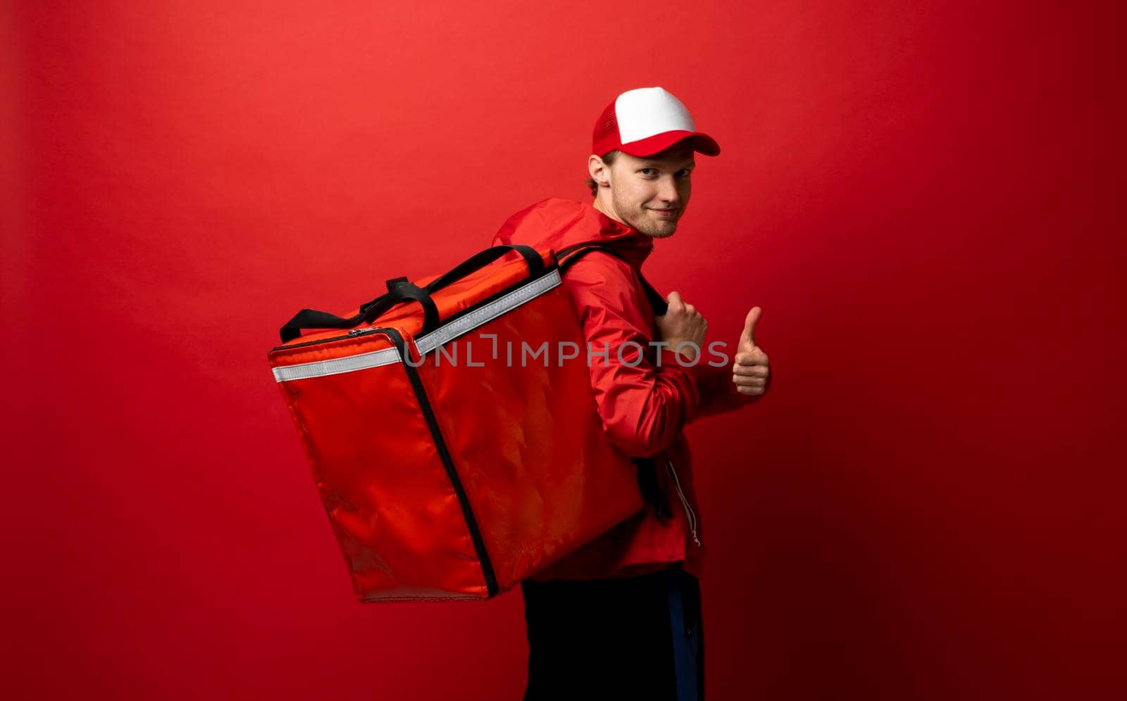 Delivery guy employee man in red cap and uniform workwear work as dealer courier hold red thermal food bag and showing thumb up isolated on red color background studio. Service concept