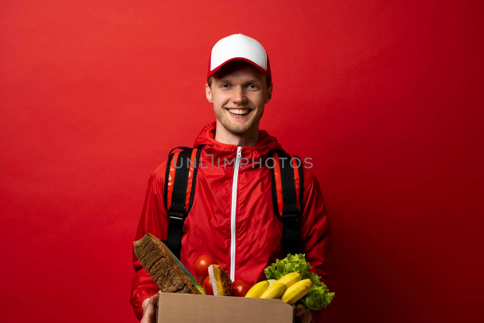 Delivery man in a red uniform with a thermal bag on a shoulder delivering food to a customer at home. Online grocery shopping service concept