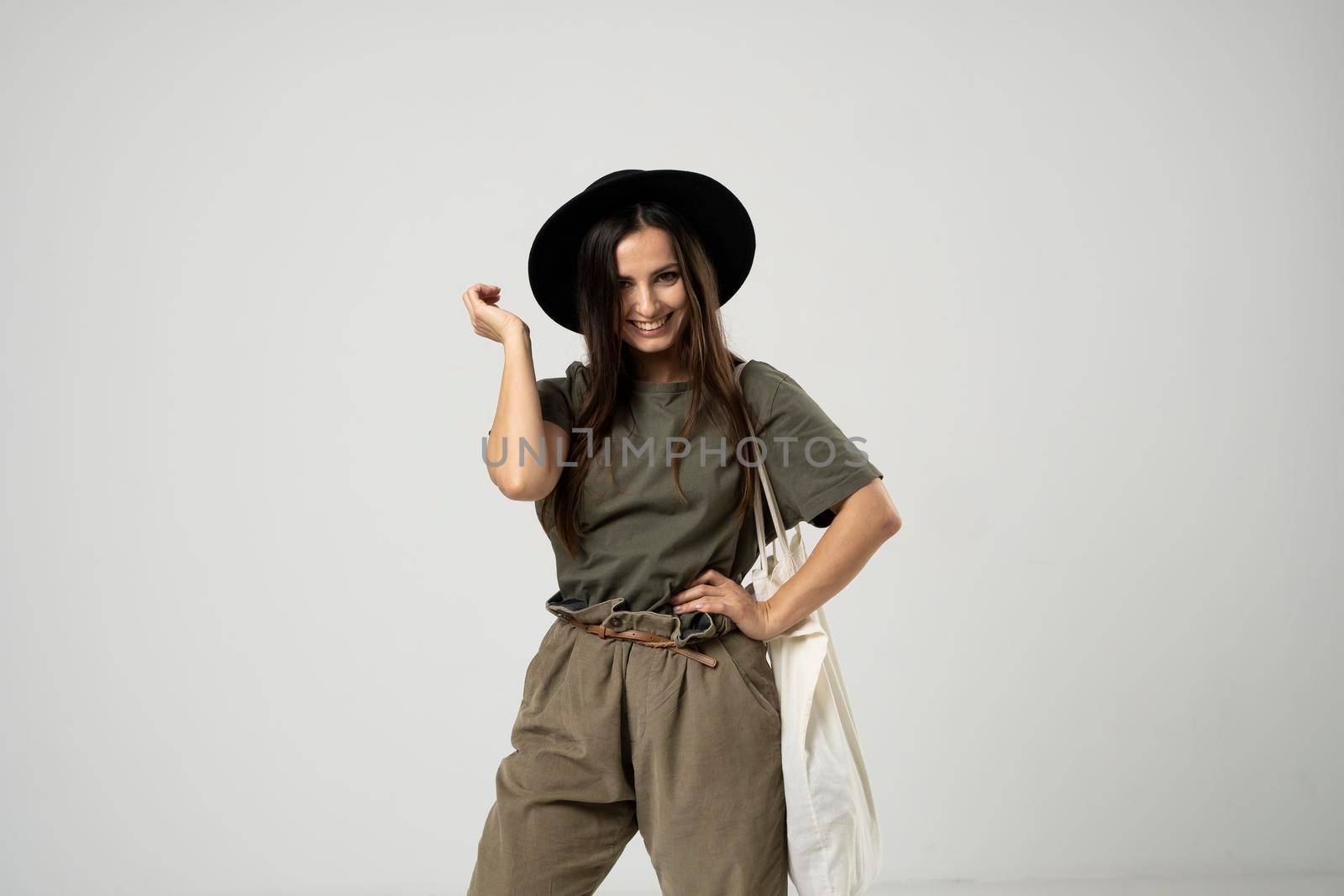 Brunette woman in green t-shirt and black hat holding cotton shopper bag with vegetables, products in white room. Eco friendly shopping bags. Zero waste, plastic free concept