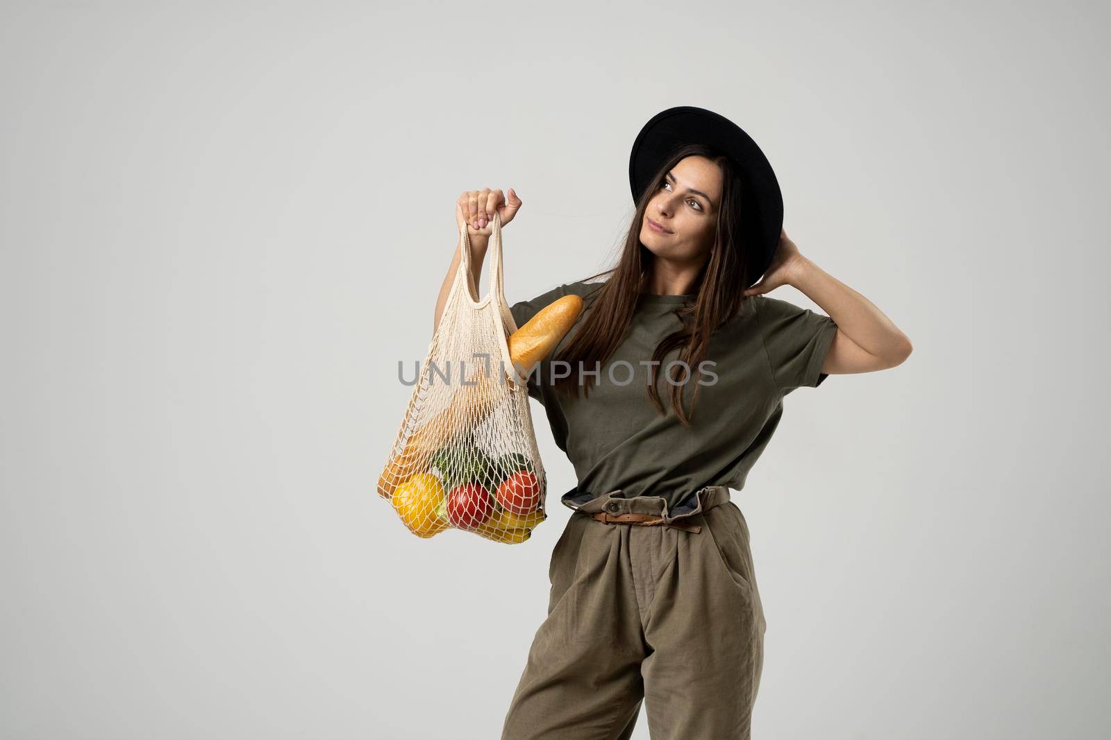 A young fashion brunete girl in a beige t-shirt and black hat holds a mesh reusable eco bag with food, vegetabes, fruits. Zero waste, plastic free. Eco friendly concept. Sustainable lifestyle