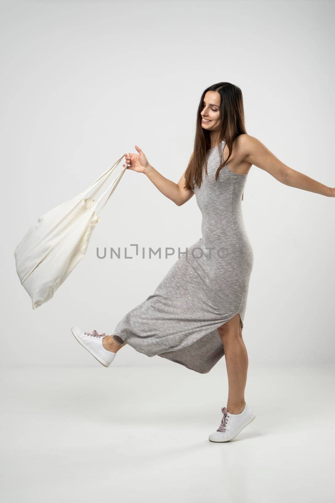 Happy young brunette woman in a grey dress dancing with a cotton bag with groceries. Zero waste concept. Eco friendly lifestyle. Mockup
