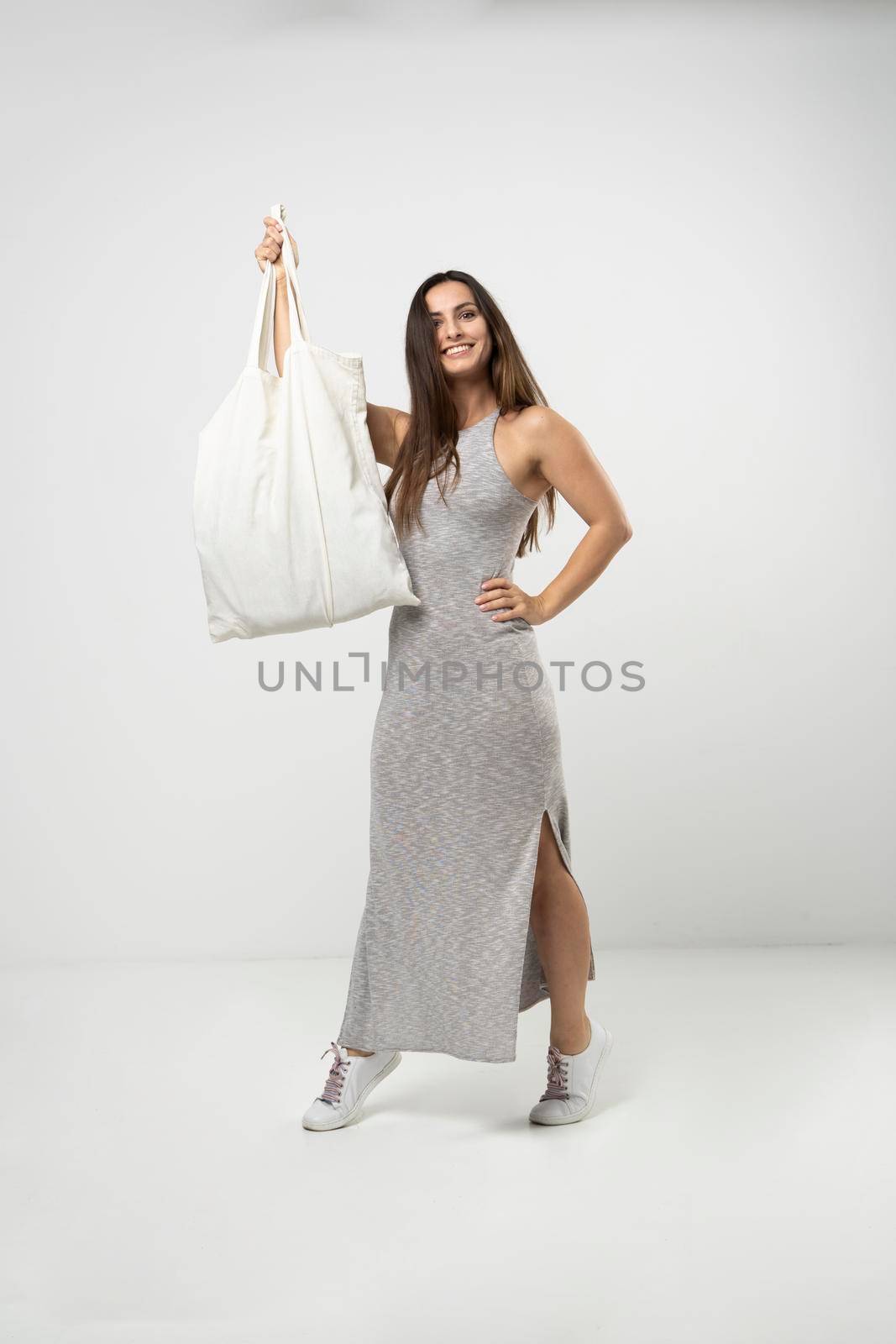 Brunette woman in grey dress holding cotton shopper bag with vegetables, products in white room. Eco friendly shopping bags. Zero waste, plastic free concept. by vovsht