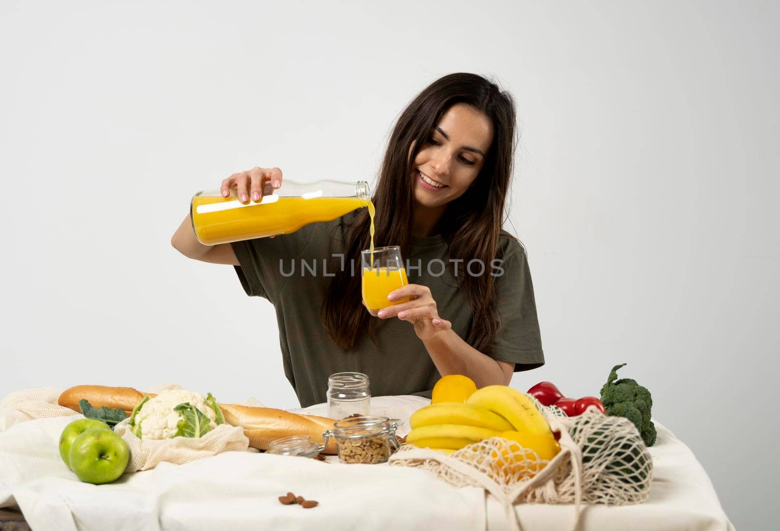 Woman in green t-shirt pouring a juice from a glass bottle in a glass over a table with mesh eco bag, healthy vegan vegetables, fruits, bread, snacks. Zero waste concept