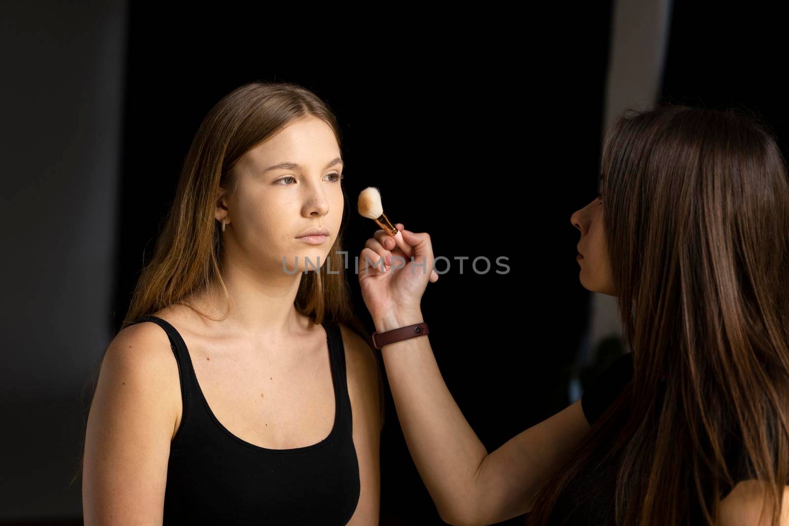 Professional make-up artist working with brush on model face. Process of making makeup. Portrait of young blonde woman in beauty salon interior. Applying tone to skin