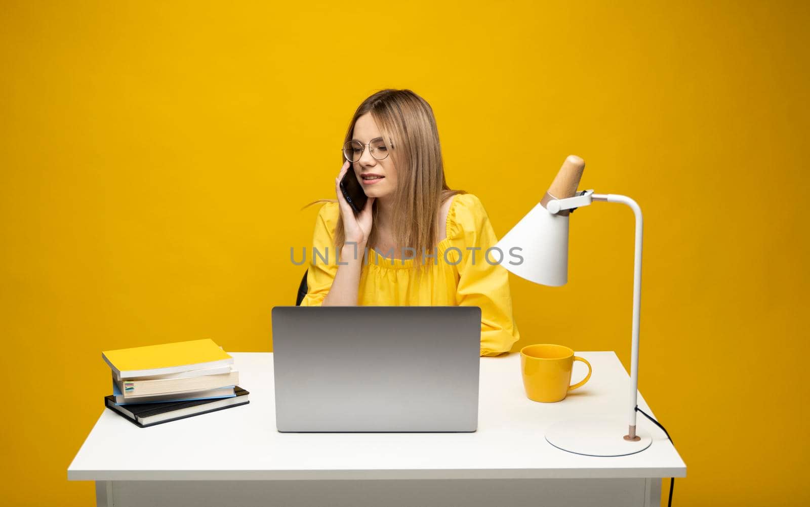 Young attractive business woman with a blond hair sitting at the table and working on a laptop and talking with a client on the phone. Young cheerful student girl talking with a smartphone. Studying. by vovsht