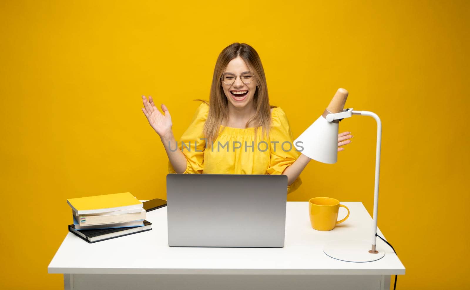 Excited young woman working with a grey laptop computer, notebook while sitting at the table with grey laptop. Smiling business woman or student received a good news isolated on a yellow background. by vovsht