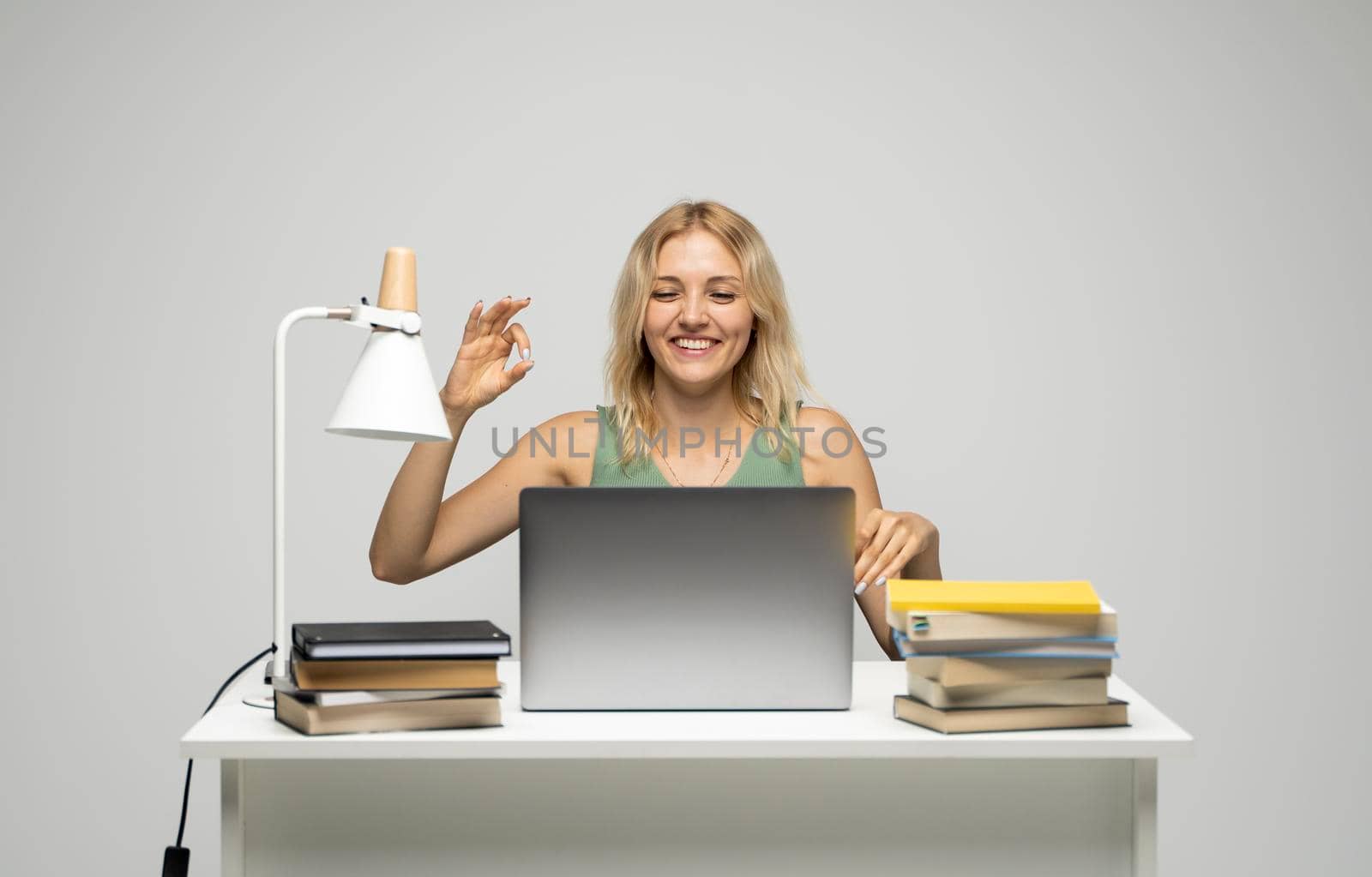 Student girl showing ok gesture with both hands, happy with completed work, smiling and looking at the camera while working on laptop computer sitting in front of big piles of books. by vovsht