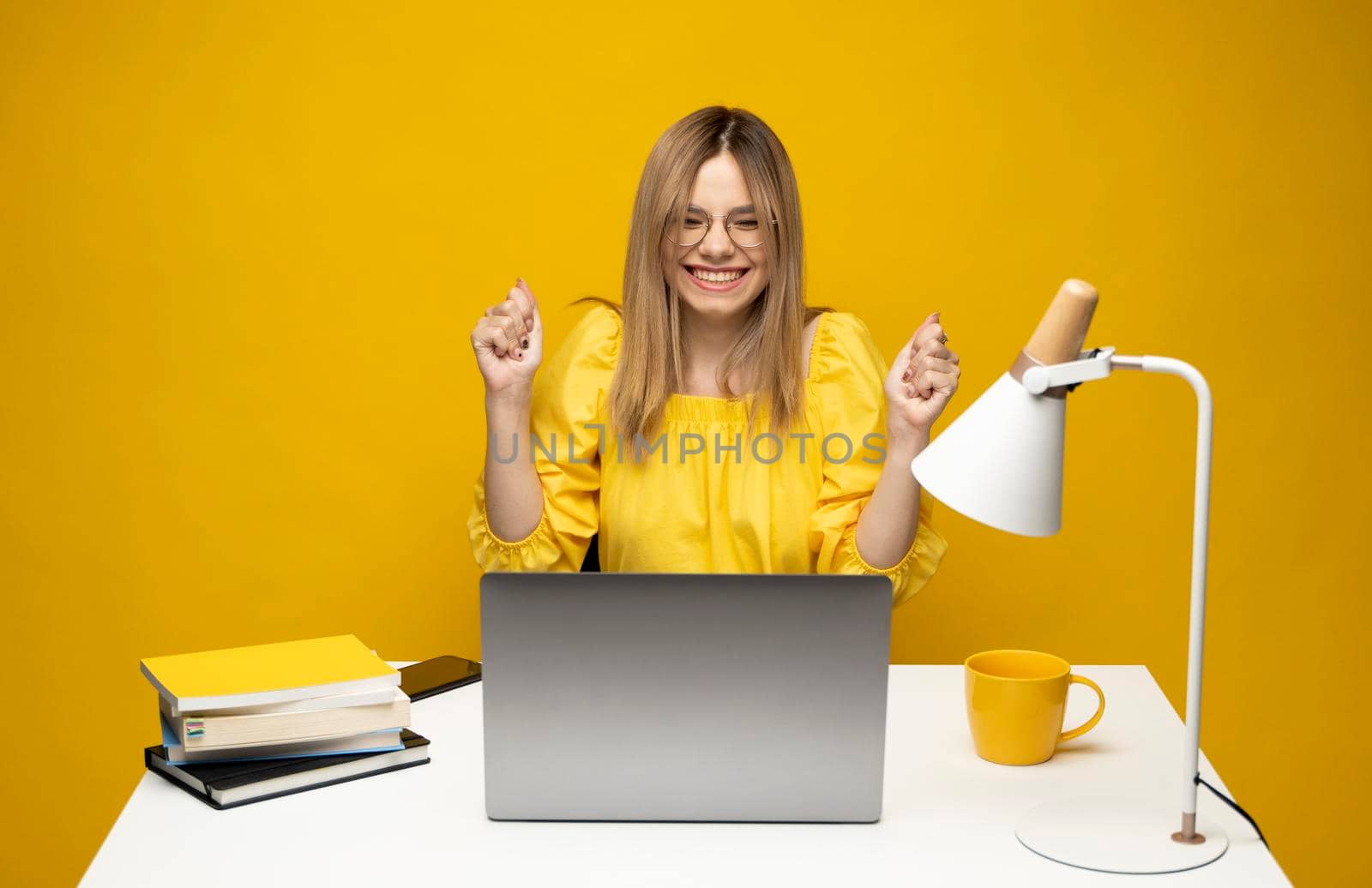 Excited young woman working with a grey laptop computer, notebook while sitting at the table with grey laptop. Smiling business woman or student received a good news isolated on a yellow background. by vovsht